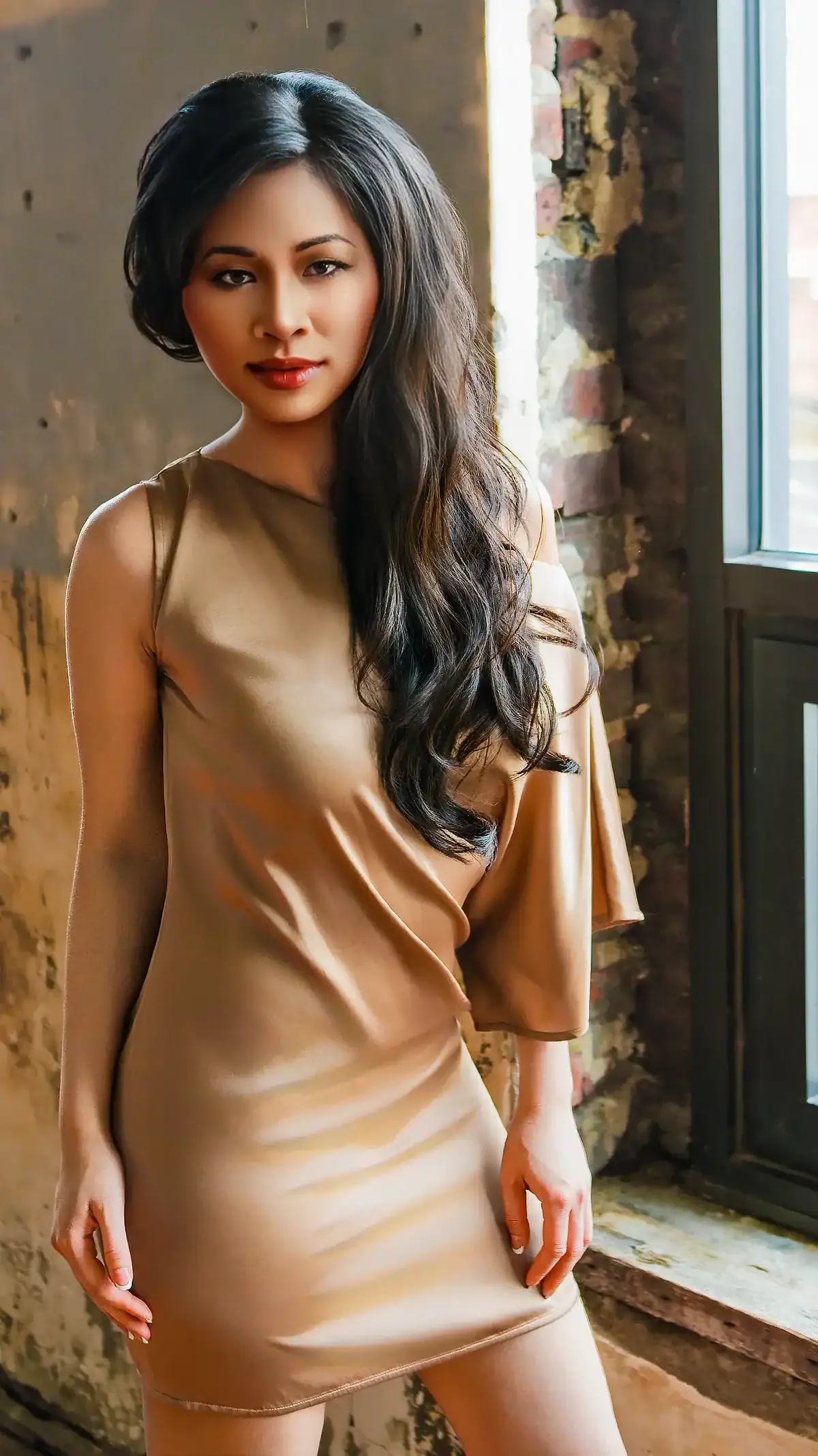 Stylish woman with long dark hair in a beige dress near a window.